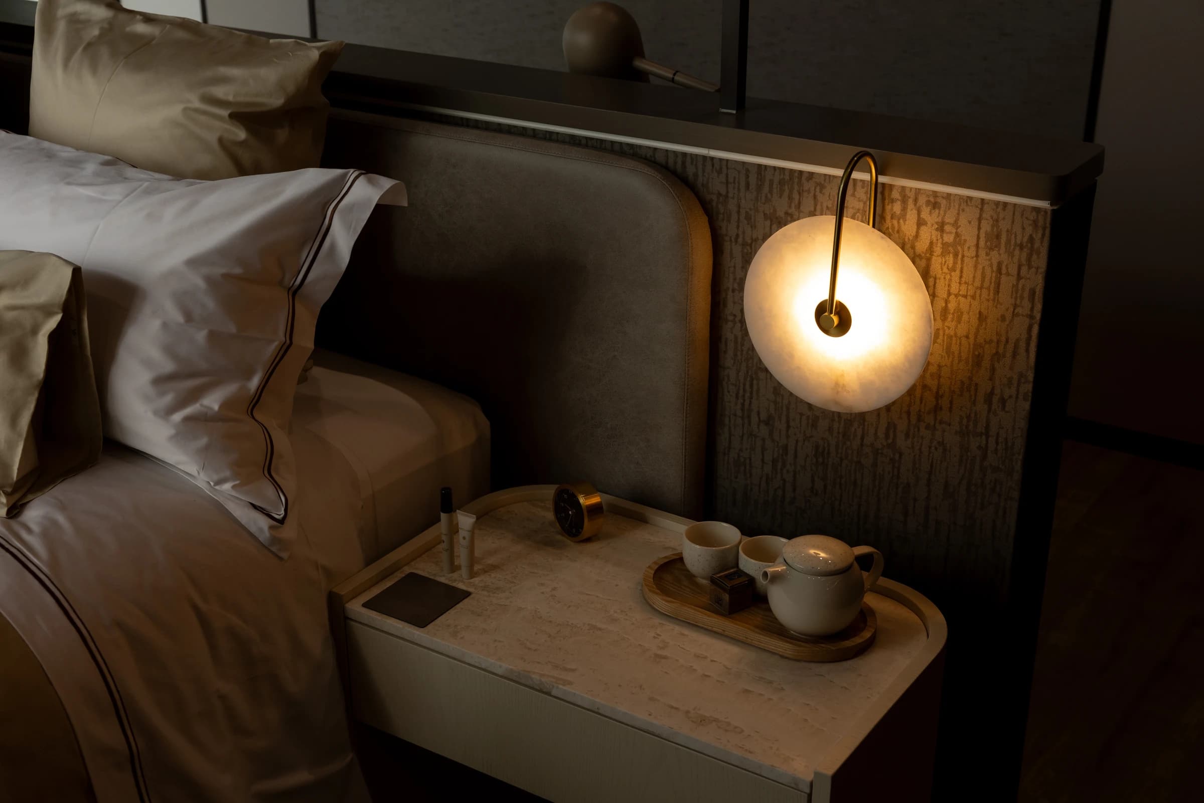 A hotel bedside with a glowing wall light, tea tray and pressed white linen